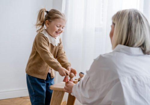 laughing child playing with wooden figurines near blurred mom at home